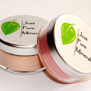 May include: Two small, round jars of mineral makeup. The jars have silver lids with a green leaf logo and the text "Just Pure Minerals." One jar contains a light peach powder, and the other contains a pink powder.