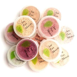 May include: A collection of small, round, clear plastic containers filled with various shades of loose mineral powder. Each lid features a green leaf graphic and the text Just Pure Minerals printed in a dark, simple, sans-serif font.