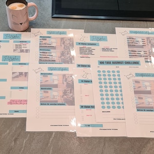 Könnte beinhalten: Eine Sammlung von gedruckten Planer-Blättern mit deutschem Text, darunter "Putzplan" und "100 TAGE AUSMIST-CHALLENGE". Die Blätter sind weiß mit blauen und rosa Akzenten, und eine Tasse mit Katzen-Gesicht mit Getränk steht daneben.