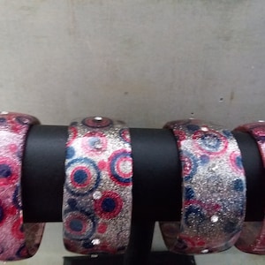 Può includere: Quattro braccialetti spessi con finitura glitterata, esposti su un supporto nero. I braccialetti presentano un motivo di cerchi rossi, blu e argentati, impreziositi da piccoli strass trasparenti. Il tema generale è la bigiotteria.