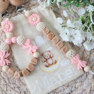 May include: A baby's pacifier clip and matching bag. The clip has pink flower and bow details, wooden beads spelling "MABEL", and a wooden clip. The bag features a teddy bear illustration and the name "Mabel".