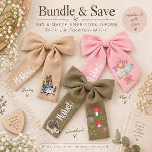 May include: Three fabric hair bows in tan, pink, and olive green. Each bow features white embroidered text reading Mabel, with additional embroidery of a rabbit, a floral bouquet, and woodland elements including a hedgehog and mushrooms.