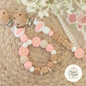 May include: Two baby pacifier clips with wooden beads spelling a name, pink and white beads, and flamingo and flower accents. The clips have wooden round clips. The background is a woven mat with floral accents.