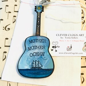 May include: A wooden guitar ornament painted with a blue ocean scene and the words "Mother Mother Ocean". The ornament is packaged in a white drawstring bag with a tag that reads "Clever Clogs Art by: Tonia Sellers Music + Art = A Happy Heart www.CleverClogsArt.com".