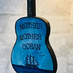 May include: A blue wooden guitar with the words "Mother Ocean" painted on the body. A small sailboat is painted on the body of the guitar.