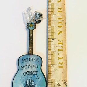 May include: A wooden guitar ornament painted with a blue ocean scene and the words "Mother Mother Ocean". The guitar has a silver bell charm attached to the top.