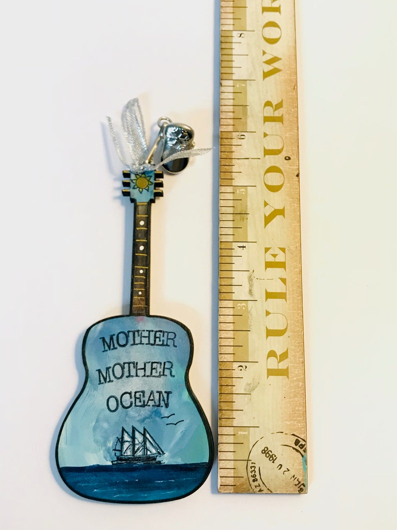 May include: A wooden guitar ornament painted with a blue ocean scene and the words "Mother Mother Ocean". The guitar has a silver bell charm attached to the top.