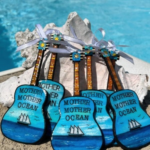 May include: Six wooden guitar ornaments painted in a blue ocean theme with white sailboats and the words "Mother Ocean" on each.