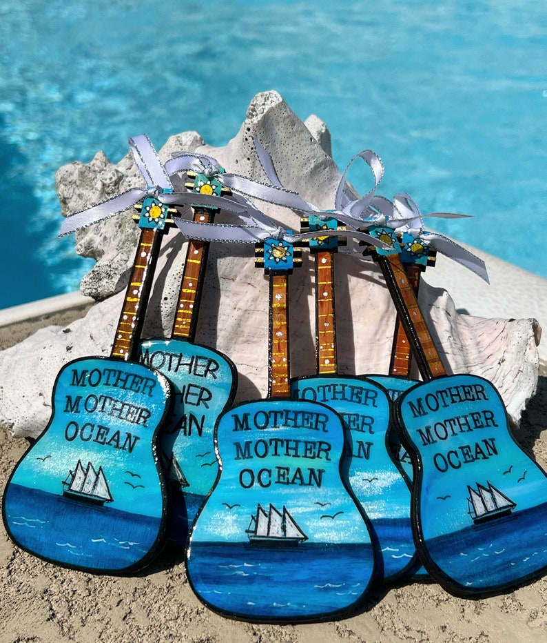 May include: Six wooden guitar ornaments painted in a blue ocean theme with white sailboats and the words "Mother Ocean" on each.