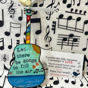 May include: A blue and green guitar-shaped ornament with the text "Let there be songs to fill the air" and a sun and hearts painted on it. The ornament is hanging from a white ribbon against a background of black musical notes.