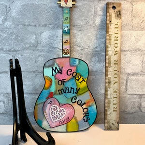 May include: A colorful wooden guitar with the words "My coat of many colors" and a pink heart with the word "DOLLY" on it. The guitar is on a black stand.