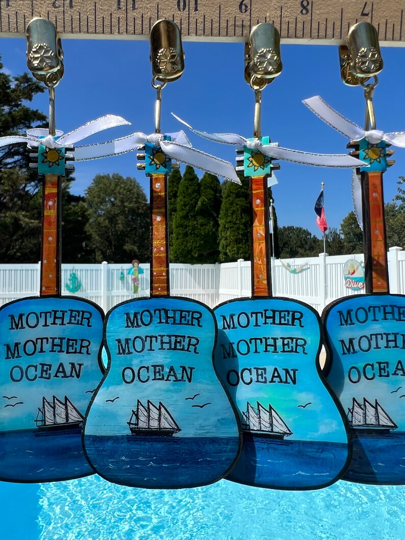 May include: Four blue and white painted wooden guitar ornaments with the text "Mother Mother Ocean" on each. The ornaments are hanging from gold clips with white ribbon.