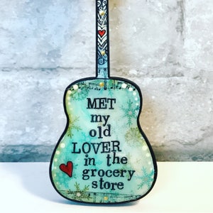 May include: A teal blue guitar-shaped sign with the text "MET my old LOVER in the grocery store" and a red heart. The guitar has a black outline and a white and black patterned neck.