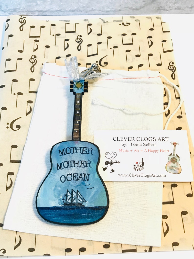 May include: A wooden guitar ornament painted with a blue ocean scene and the words "Mother Mother Ocean". The ornament is packaged in a white drawstring bag with a tag that reads "Clever Clogs Art by: Tonia Sellers Music + Art = A Happy Heart www.CleverClogsArt.com".