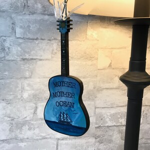 May include: A wooden guitar ornament painted blue with a sailboat and the words "Mother Mother Ocean" on the front.