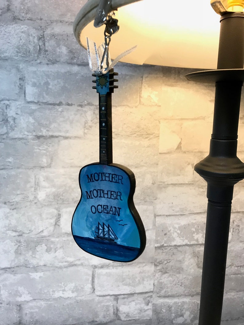 May include: A wooden guitar ornament painted blue with a sailboat and the words "Mother Mother Ocean" on the front.