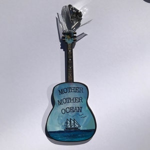 May include: A wooden guitar ornament painted with a blue ocean scene and the words "Mother Mother Ocean". A small sailboat is painted on the ocean with two birds flying above it.