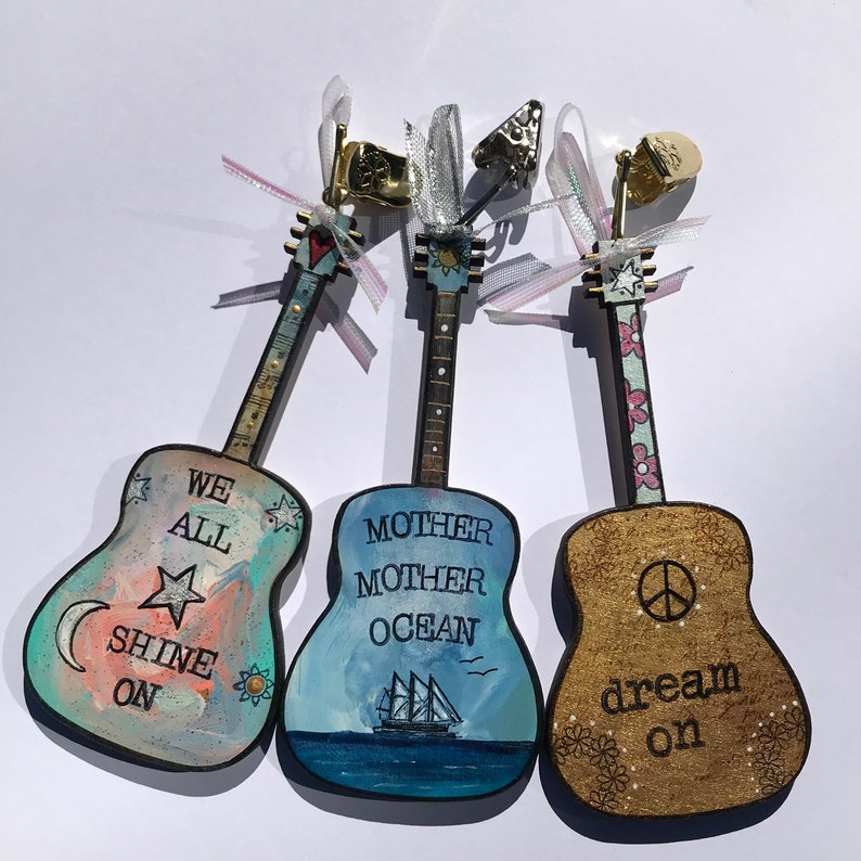 May include: Three wooden guitar ornaments with painted designs. The first guitar is blue and pink with the text "We All Shine On". The second guitar is blue with the text "Mother Mother Ocean". The third guitar is brown with the text "dream on" and a peace sign.