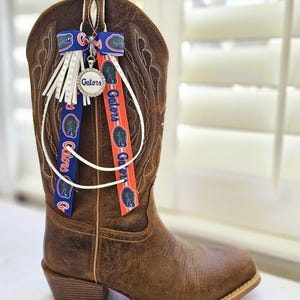 Puede incluir: Una bota de vaquero de cuero marr&oacute;n con costuras decorativas. La bota est&aacute; adornada con cintas azules y naranjas con la palabra "Gators" y un dije con la palabra "Gators" en un c&iacute;rculo. Flecos blancos cuelgan de la parte superior.