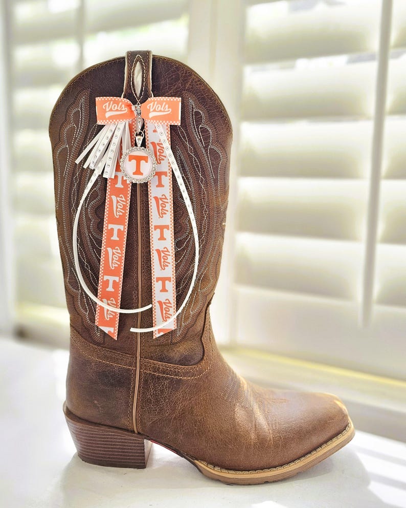 May include: Brown leather cowboy boot with decorative orange and white ribbons. The ribbons feature the word "Vols" and a "T" logo. A silver charm with a "T" logo hangs from the boot.