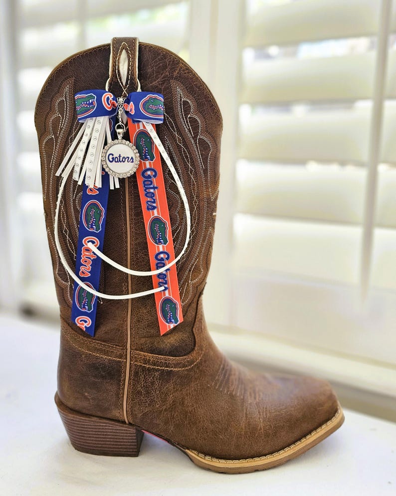 Puede incluir: Una bota de vaquero de cuero marr&oacute;n con costuras decorativas. La bota est&aacute; adornada con cintas azules y naranjas con la palabra "Gators" y un dije con la palabra "Gators" en un c&iacute;rculo. Flecos blancos cuelgan de la parte superior.