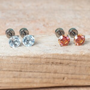 May include: Two pairs of stud earrings featuring round-cut stones. One pair displays light blue stones, while the other pair features orange-toned stones. Each earring has a metallic post and a circular backing, showcasing a simple, classic design.