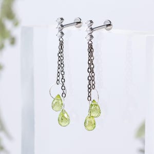 Puede incluir: Un par de pendientes plateados con un poste y tres piedras transparentes. Los pendientes tienen una cadena que se conecta a dos piedras en forma de lágrima de color verde claro. Los pendientes se muestran sobre una superficie blanca.
