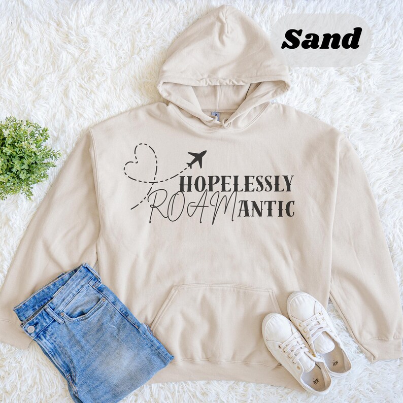 May include: A sand-colored hooded sweatshirt with the words "HOPLESSLY ROAMANTIC" printed in black. The design includes a heart-shaped dotted line and a small airplane graphic. The sweatshirt is paired with blue jeans and white sneakers.