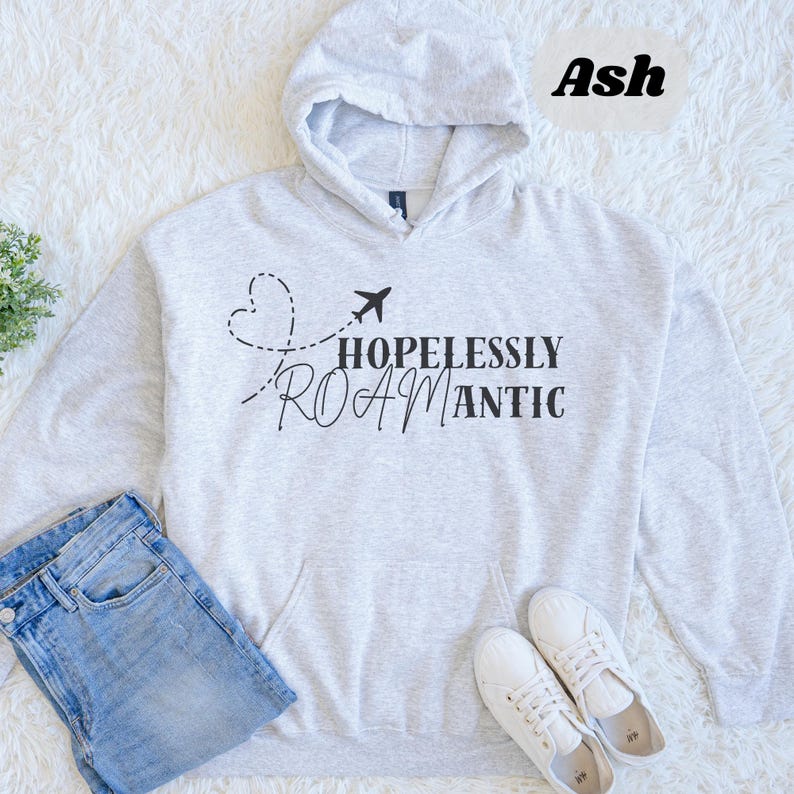 May include: A light grey hoodie with the text "HOPELESSLY ROAMANTIC" in black, featuring a dotted heart and airplane graphic. The hoodie has a front pocket and a hood. A pair of blue jeans and white trainers are also in the image.