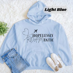May include: Light blue hoodie with the text "HOPLESSLY ROAMANTIC" and a graphic of a heart and airplane. The hoodie has a hood and a front pocket. The words "Light Blue" are in a white box in the upper right corner.