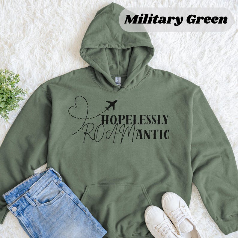 May include: Military green hoodie with a front pocket and a hood. The hoodie features the text "HOPELESSLY ROAMANTIC" with a dotted heart and airplane graphic. A pair of blue jeans and white sneakers are visible.