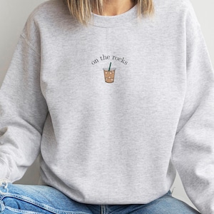 May include: A light gray sweatshirt with the phrase "on the rocks" above a graphic of an iced coffee drink. The sweatshirt is a crew neck style and is made of a soft material. The model is wearing blue jeans.
