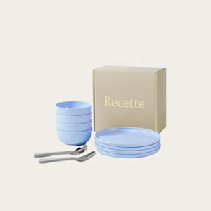 May include: A light blue dinnerware set includes stacked bowls, plates, forks, spoons, and a box with the word "Recette" printed on it. The plates and bowls are stacked neatly. The utensils are silver.