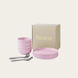 May include: A pink dinnerware set includes stacked bowls, plates, and forks. The set is packaged in a beige box with the word "Recette" printed on the front. The plates and bowls are a soft pink color.