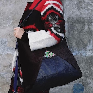 May include: A black tote bag with a blue bottom section is carried over the shoulder. The bag has a decorative patch with green, pink, and blue embroidery. The person is wearing a red, black, and white patterned coat.