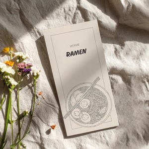 Könnte beinhalten: Eine beige Karte mit den Worten "LET'S EAT RAMEN" und einer Strichzeichnung einer Ramen-Schüssel mit Essstäbchen. Die Karte liegt auf einer strukturierten, cremefarbenen Oberfläche, mit einem kleinen Blumenstrauß links.