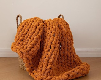 May include: A chunky knit blanket in a warm orange hue, draped over a woven basket. The blanket's thick, textured weave is visible, suggesting a cozy and comfortable item. The basket has two handles. The blanket is a home decor item.
