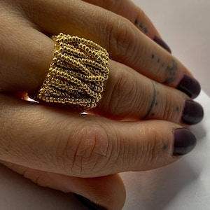 Pode incluir: Um anel dourado com um design de contas. O anel é largo e cobre uma parte significativa do dedo. As pequenas contas estão muito juntas, criando uma superfície texturizada. O anel é de cor dourada brilhante. Joalharia de moda.