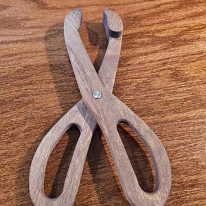 May include: Wooden tongs with a natural finish. The tongs have a scissor-like design with curved handles and a small opening at the end for gripping. The wood grain is visible, and a metal rivet secures the two pieces together.