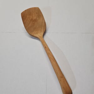 Puede incluir: Una espátula de madera con una cabeza ancha y plana y un mango largo y redondeado. La espátula está hecha de madera de color claro, mostrando la veta natural. El mango tiene un pequeño agujero cerca del extremo. Este utensilio de cocina es ideal para cocinar y servir.