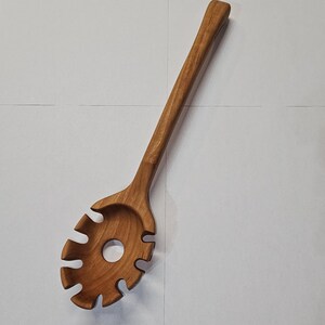 Puede incluir: Una cuchara de espagueti de madera con un mango largo y una cabeza ovalada ranurada. La cuchara está hecha de madera de color marrón claro y tiene un acabado liso y pulido. El diseño incluye un agujero central y ranuras curvas para escurrir la pasta.