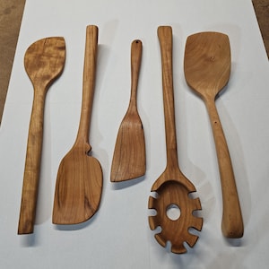 May include: Five wooden kitchen utensils, including a spoon, spatula, and slotted spoon, are arranged on a white surface. The utensils are made of light brown wood with visible grain patterns. The handles are long and slender, and the tools have various shapes for different cooking tasks.