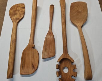 May include: Five wooden kitchen utensils, including a spoon, spatula, and slotted spoon, are arranged on a white surface. The utensils are made of light brown wood with visible grain patterns. The handles are long and slender, and the tools have various shapes for different cooking tasks.