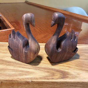 Könnte beinhalten: Zwei handgefertigte Schwanfiguren aus Holz. Die Schwäne sind aus dunkelbraunem Holz geschnitzt, mit detaillierten Flügeln und gebogenen Hälsen. Sie werden auf einer Holzoberfläche ausgestellt, die die natürliche Maserung des Holzes zeigt. Die Schwäne sind ein dekorativer Gegenstand.