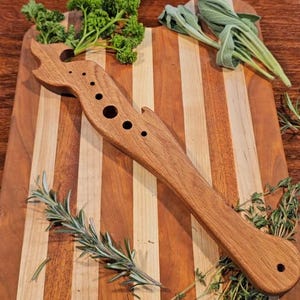 Puede incluir: Una tabla de cortar de madera a rayas con un picador de hierbas de madera. Hierbas frescas, como perejil, salvia, romero y tomillo, están dispuestas alrededor de la tabla. El picador tiene un diseño único con varios agujeros.