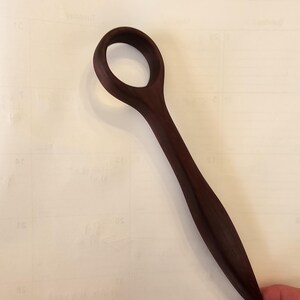 May include: A dark brown wooden spoon with a circular handle and a long, slender stem. The spoon is made of a single piece of wood and has a smooth, polished finish. The spoon is held by a hand.