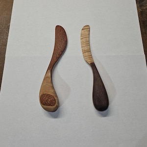 Puede incluir: Dos untadores de mantequilla de madera. Uno es de color marrón claro con un diseño tallado en el mango. El otro es de color marrón oscuro con un patrón rayado en la hoja. Ambos tienen mangos y hojas curvados.