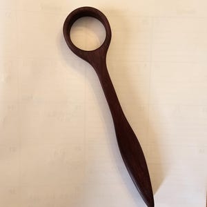 May include: A handcrafted wooden spoon with a dark brown finish. The spoon features a circular handle and a long, slender body. The spoon is made of a solid piece of wood and has a smooth, polished surface.