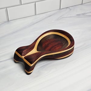 May include: A spoon rest crafted from dark brown and light tan wood, featuring a smooth, polished finish and a distinct, curved shape with a wide, rounded bowl and a tapered handle end, showcasing natural wood grain patterns throughout.
