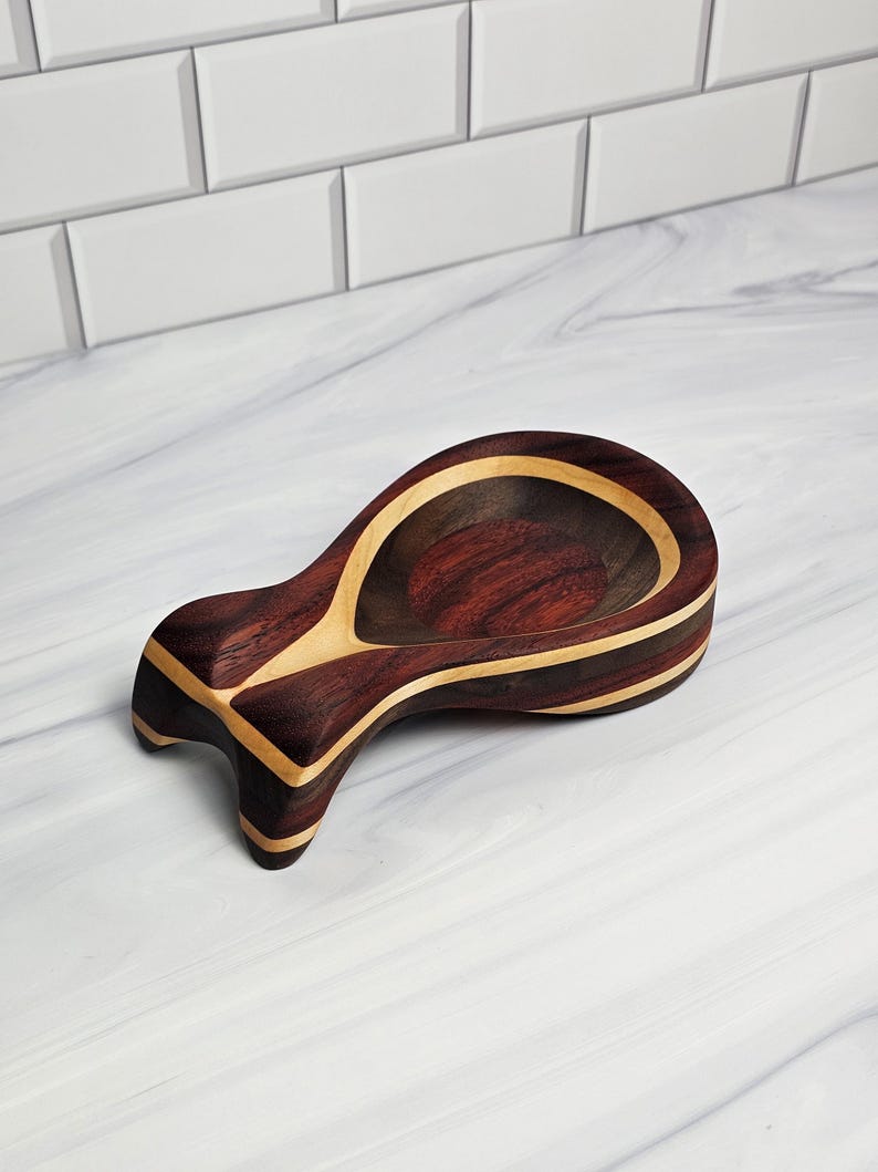 May include: A spoon rest crafted from dark brown and light tan wood, featuring a smooth, polished finish and a distinct, curved shape with a wide, rounded bowl and a tapered handle end, showcasing natural wood grain patterns throughout.